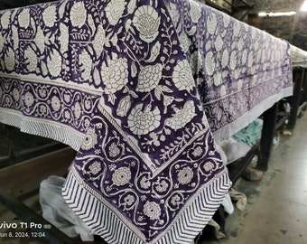 Puede incluir: Un mantel de mesa de algodón blanco con estampado floral morado. El mantel tiene un borde con un patrón repetitivo de flores y hojas blancas sobre un fondo morado.