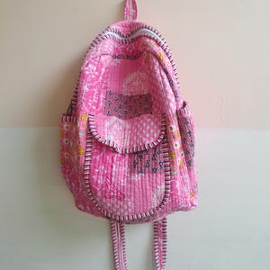 May include: A quilted backpack featuring a patchwork design in shades of pink, white, and yellow with floral and geometric patterns. The bag includes a front pocket with a flap, side pockets, and black and white striped trim along the edges.