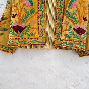 May include: Mustard yellow embroidered jacket with floral and leaf patterns in pink, green, and burgundy. The jacket has a striped lining and detailed embroidery, showcasing a bohemian style.