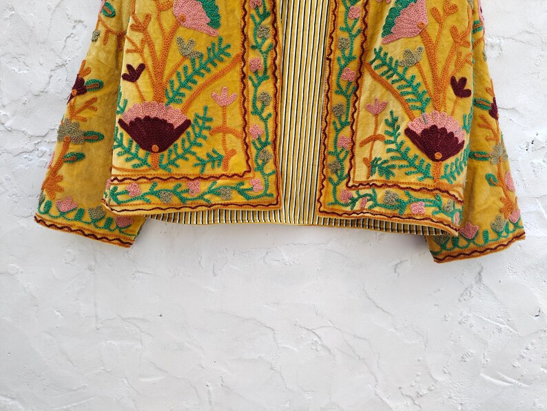 May include: Mustard yellow embroidered jacket with floral and leaf patterns in pink, green, and burgundy. The jacket has a striped lining and detailed embroidery, showcasing a bohemian style.