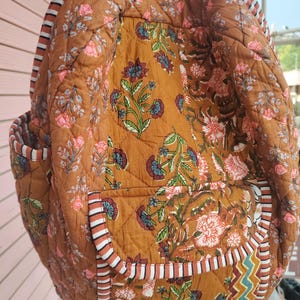 May include: A quilted backpack in a warm brown colour, decorated with a floral pattern in pink, blue, and green. The bag features striped trim and multiple pockets, offering a blend of style and functionality. The backpack is designed for carrying essentials.