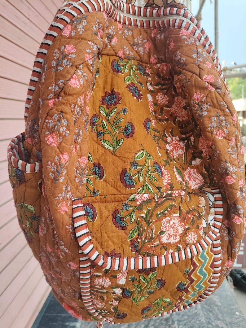 May include: A quilted backpack in a warm brown colour, decorated with a floral pattern in pink, blue, and green. The bag features striped trim and multiple pockets, offering a blend of style and functionality. The backpack is designed for carrying essentials.