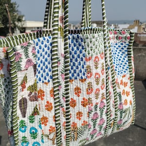 May include: A quilted tote bag with a patchwork design. The bag features a variety of floral and geometric patterns in shades of blue, orange, pink, and green on a white background. The handles are striped green and white.