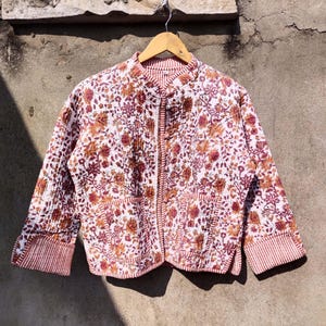 Puede incluir: Chaqueta acolchada con estampado floral en tonos burdeos, naranja y blanco. Cuello mandarín, mangas largas y dos bolsillos delanteros. Los puños y el ribete tienen un diseño a rayas.
