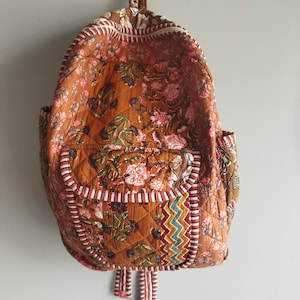 May include: A floral-patterned quilted backpack in shades of brown, pink, and green. It features a front pocket, side pockets, and adjustable straps. The edges are trimmed with white and red striped fabric.
