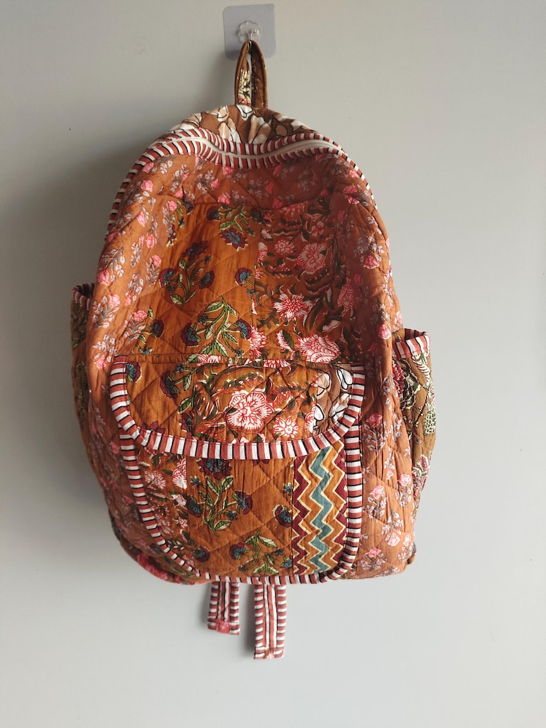 May include: A floral-patterned quilted backpack in shades of brown, pink, and green. It features a front pocket, side pockets, and adjustable straps. The edges are trimmed with white and red striped fabric.