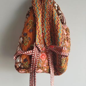 May include: Quilted backpack with a floral design in orange, brown, and white. The back of the bag has a zig-zag pattern, and it has striped ties. The bag is suspended against a plain white wall.