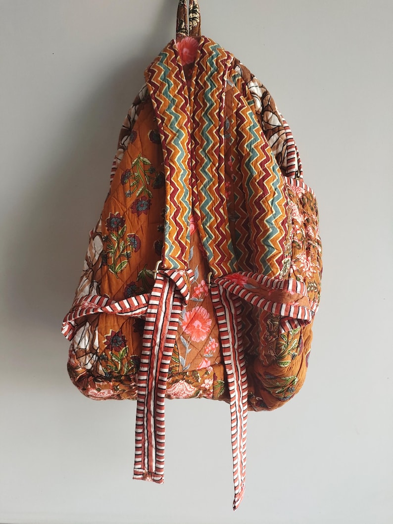 May include: Quilted backpack with a floral design in orange, brown, and white. The back of the bag has a zig-zag pattern, and it has striped ties. The bag is suspended against a plain white wall.
