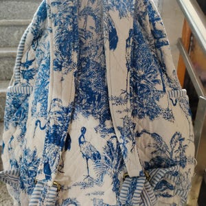 Puede incluir: Mochila acolchada con un estampado toile de Jouy azul y blanco que representa una escena tropical. El diseño incluye árboles, aves y tigres. La mochila tiene correas ajustables y bolsillos laterales con detalles a rayas.