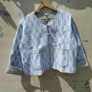 May include: White quilted jacket with a green floral pattern and two front pockets. The jacket has a round collar and is buttoned up. The cuffs and trim are green and white striped. The jacket is hanging on a wooden hanger.