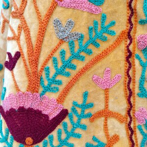 May include: Close-up of a mustard textile with detailed embroidery. The design showcases textured flowers in pink, teal, and burgundy, with orange stems and teal leaves. A burgundy border trims the edges of the fabric.