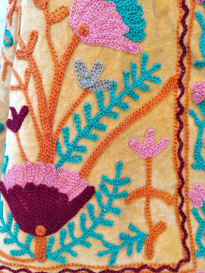 May include: Close-up of a mustard textile with detailed embroidery. The design showcases textured flowers in pink, teal, and burgundy, with orange stems and teal leaves. A burgundy border trims the edges of the fabric.