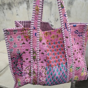 May include: A pink quilted tote bag with floral and geometric patterns. The bag has two long straps with white and black stripes.