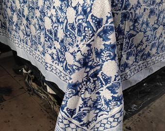 Puede incluir: Un mantel floral azul y blanco. El mantel presenta un patrón repetitivo de grandes flores y follaje estilizado en blanco sobre un fondo azul oscuro. El borde tiene un diseño a rayas y geométrico.