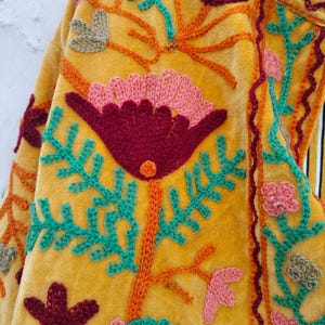 May include: Mustard yellow embroidered jacket with a floral design. The jacket features a large, central flower in deep red and pink, surrounded by teal and orange embroidered leaves and stems. Additional small flowers and leaf clusters are scattered across the fabric.
