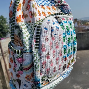 Peut inclure: Sac à dos matelassé avec un motif patchwork à motifs floraux multicolores. Il est orné de fleurs roses, oranges, vertes et bleues sur fond blanc. Il possède un compartiment principal zippé et des poches latérales.