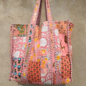 May include: A patchwork tote bag with a pink floral design. The bag features various floral patterns in shades of pink, orange, and white. The handles and edges are trimmed with a striped pattern. The bag is made of quilted fabric.