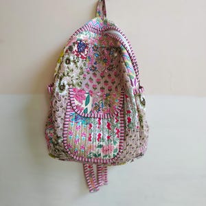 Könnte beinhalten: Ein gesteppter Rucksack mit Patchwork-Design und verschiedenen Blumenmustern in Rosa-, Grün- und Beigetönen. Die Tasche verfügt über eine Vordertasche mit Klappe, einen Tragegriff und verstellbare Schultergurte, die alle mit rosa-weiß gestreiften Kanten versehen sind.