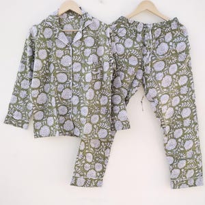 May include: A two-piece pajama set in olive green with a white floral pattern. The long-sleeved button-down shirt and matching pants have white piping along the edges. The set is displayed on wooden hangers.