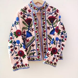 May include: A pink embroidered jacket with floral patterns in shades of red, blue, and green. The jacket has a stand-up collar and a front zipper closure.