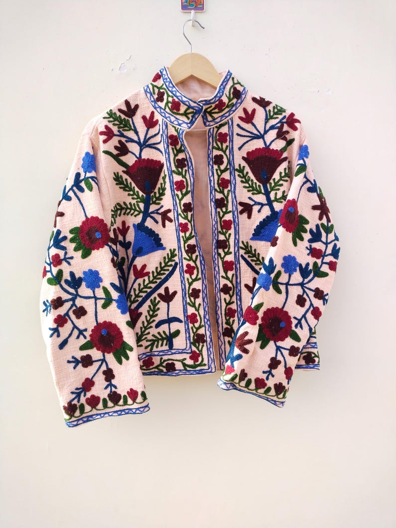 May include: A pink embroidered jacket with floral patterns in shades of red, blue, and green. The jacket has a stand-up collar and a front zipper closure.