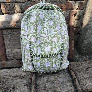 Puede incluir: Una mochila con estampado floral verde y blanco con asa superior y bolsillo delantero. La mochila tiene un diseño acolchado con ribete verde y borde a rayas blanco y negro. La bolsa está sobre una superficie de madera.