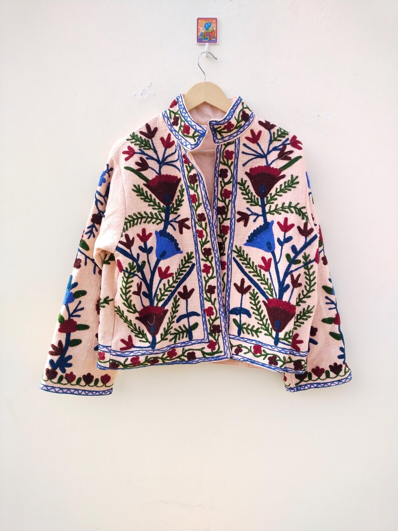 May include: A pink embroidered jacket with a floral pattern in shades of blue, green, red, and white. The jacket has a stand-up collar and a button closure.
