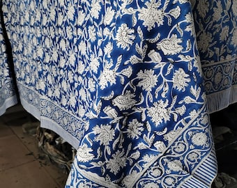 Puede incluir: Un mantel floral azul y blanco. La tela presenta un patrón repetido de flores y hojas blancas sobre un fondo azul intenso. El borde tiene un diseño floral detallado con rayas. El mantel está drapeado sobre una superficie.