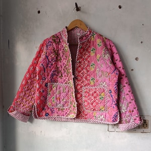 Può includere: Una giacca trapuntata rosa con stampa floreale. La giacca ha un colletto alla coreana e una chiusura con bottoni. Il tessuto è a patchwork e presenta una varietà di motivi floreali.