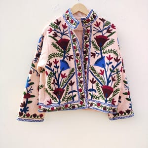 May include: A pink embroidered jacket with a floral pattern in shades of blue, green, red, and white. The jacket has a high collar and a button closure.