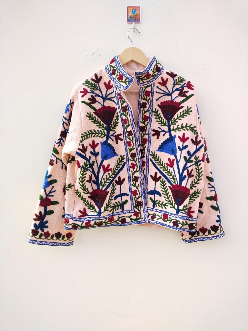 May include: A pink embroidered jacket with a floral pattern in shades of blue, green, red, and white. The jacket has a high collar and a button closure.