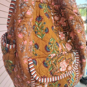 May include: A quilted backpack in a warm brown colour, decorated with floral patterns in pink, blue, and green. The bag has striped accents and pockets, offering a blend of style and functionality. The design showcases a bohemian aesthetic.