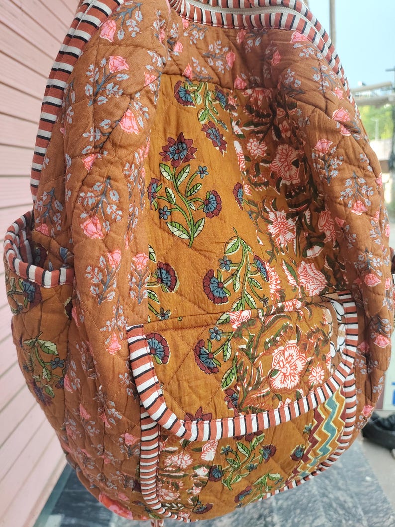 May include: A quilted backpack in a warm brown colour, decorated with floral patterns in pink, blue, and green. The bag has striped accents and pockets, offering a blend of style and functionality. The design showcases a bohemian aesthetic.