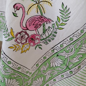 May include: White tablecloth with a green and black floral border. A pink flamingo with a yellow palm tree and pink flowers are printed in the centre of the tablecloth.