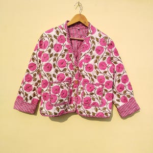 May include: A quilted jacket with a floral pattern in pink and brown on a white background. The jacket has a V-neck, two pockets, and is lined with a pink and white striped fabric. It is hanging on a wooden hanger.
