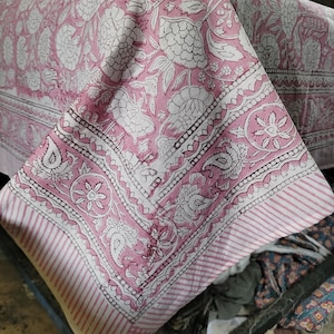 May include: Pink cotton tablecloth with a white floral print and a striped border. The tablecloth is folded in half and the corner is visible.