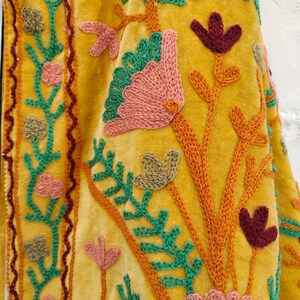 May include: Close-up of a mustard yellow textile with intricate floral embroidery. The embroidery features pink, green, and burgundy flowers and leaves, with orange stems and vines. The fabric has a soft, velvety texture.