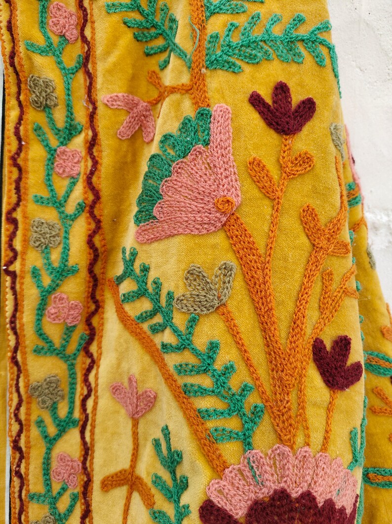 May include: Close-up of a mustard yellow textile with intricate floral embroidery. The embroidery features pink, green, and burgundy flowers and leaves, with orange stems and vines. The fabric has a soft, velvety texture.