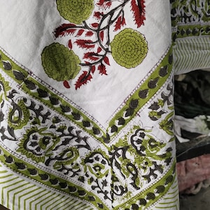 May include: A white cotton tablecloth with a green and black floral print. The tablecloth has a border of green and black paisley designs. The tablecloth is folded in half and the corner is visible.