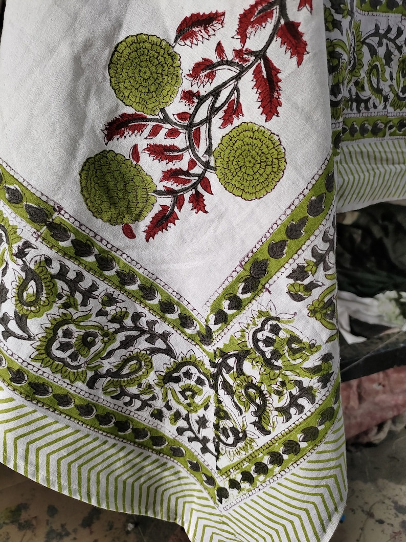 May include: A white cotton tablecloth with a green and black floral print. The tablecloth has a border of green and black paisley designs. The tablecloth is folded in half and the corner is visible.