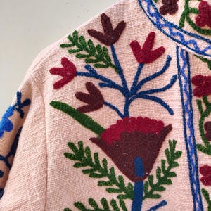 May include: A pink fabric jacket with colorful embroidered flowers. The flowers are red, green, blue, and brown. The jacket has a high collar and a button closure.