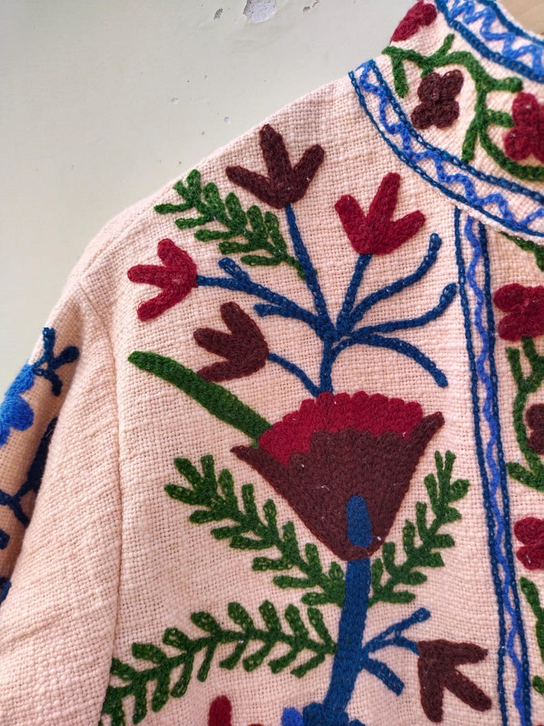 May include: A pink fabric jacket with colorful embroidered flowers. The flowers are red, green, blue, and brown. The jacket has a high collar and a button closure.