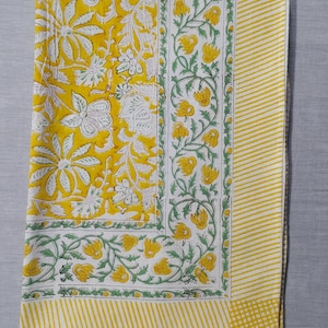 May include: A yellow and white floral patterned tablecloth. The tablecloth features a large floral design in yellow and white, with a border of yellow and white stripes. The edges are trimmed with a green and white floral pattern.