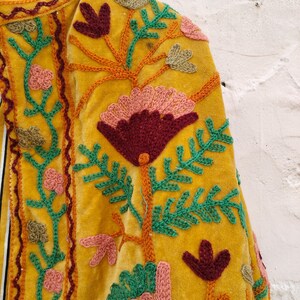 May include: Close-up of a mustard yellow velvet jacket with detailed floral embroidery. The embroidery uses burgundy, pink, green, and orange thread, forming a botanical design. The jacket's edges are trimmed with a decorative stitch.