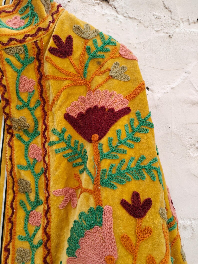 May include: Close-up of a mustard yellow velvet jacket with detailed floral embroidery. The embroidery uses burgundy, pink, green, and orange thread, forming a botanical design. The jacket's edges are trimmed with a decorative stitch.