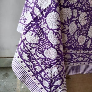 May include: Purple and white cotton tablecloth with a floral print. The tablecloth has a border with a striped pattern.