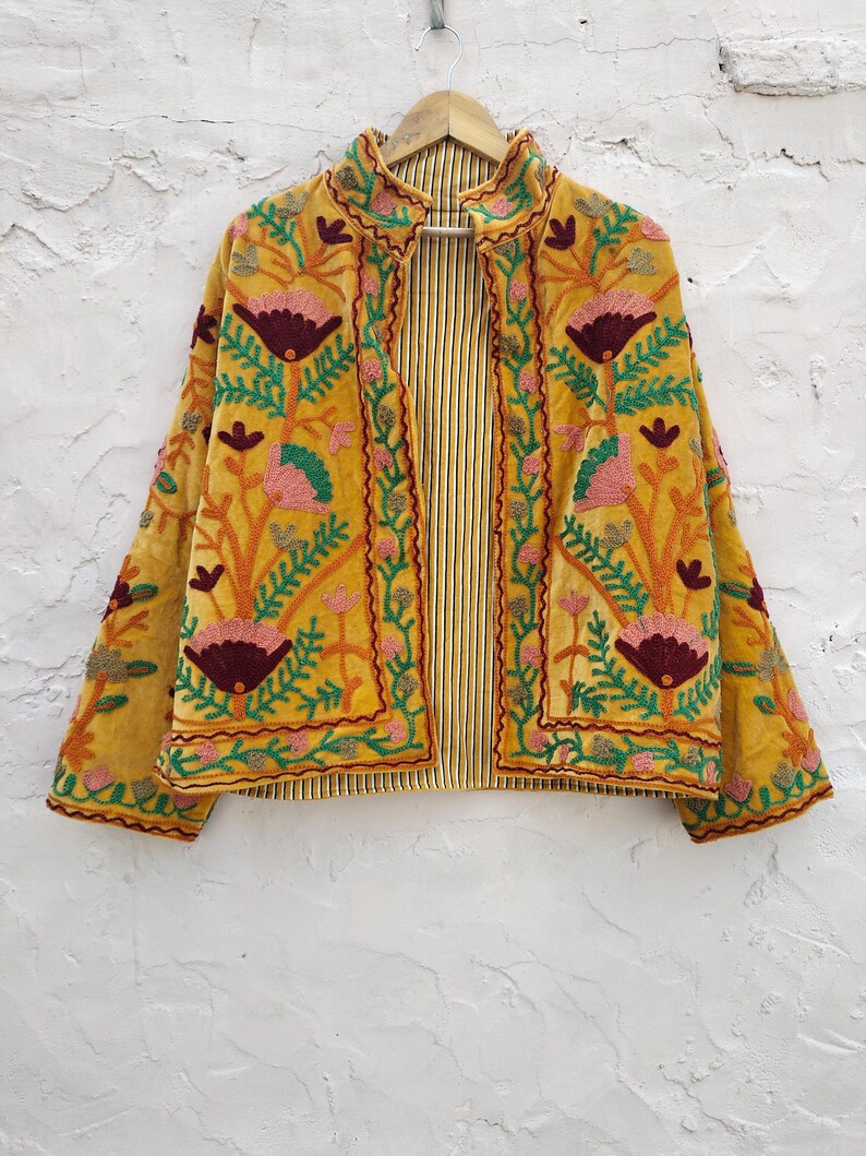May include: Mustard yellow embroidered jacket with floral and leaf patterns in pink, green, and burgundy. The jacket has a striped lining and is hanging on a wooden hanger. The jacket's design is a blend of colours and textures.