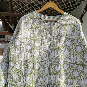 May include: A reversible quilted jacket with a floral pattern in shades of green and white. The jacket has a button-up front and a striped lining. The jacket is hanging on a wooden hanger.