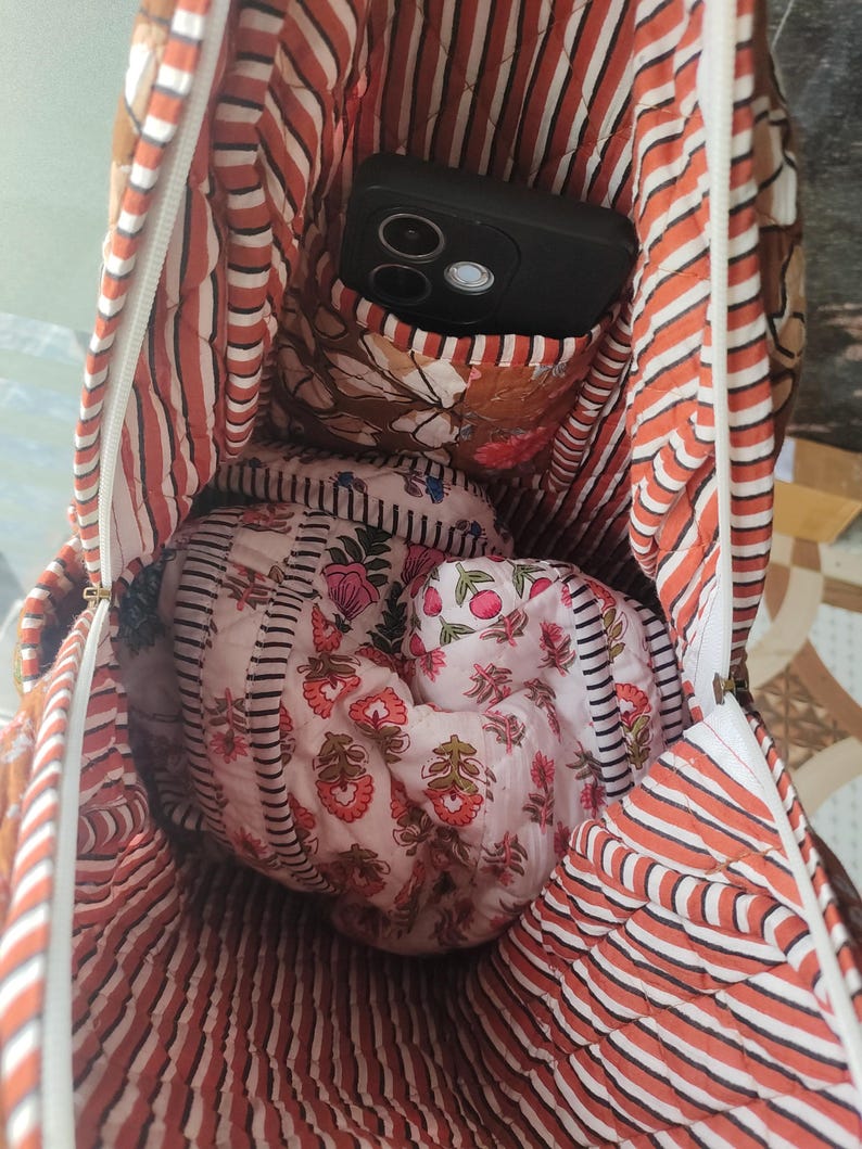 May include: Inside view of a quilted tote bag with a black phone in the pocket. The bag has a striped pattern in orange and white, with floral fabric inside. A smaller, floral-printed pouch is also visible within the bag's interior.