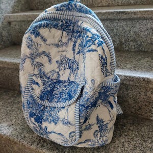 May include: A quilted backpack featuring a blue and white toile pattern with a jungle scene. The backpack has a top handle, two side pockets, and a striped blue trim. The main pattern includes tigers, palm trees, and other tropical elements.